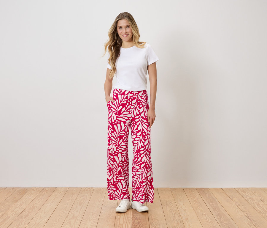 Une femme portant un t-shirt blanc et un pantalon rouge et blanc à imprimé floral se tient devant un mur, une main dans la poche.