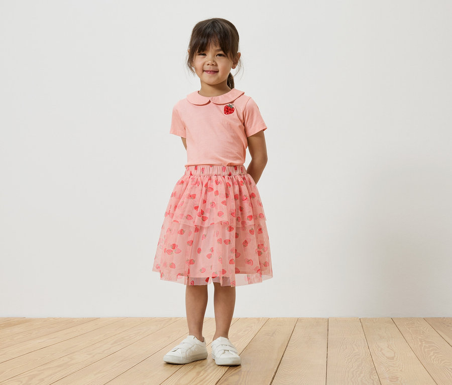 Une jeune fille se tient devant un mur blanc, portant un t-shirt rose avec un motif de fraise, une jupe en tulle rose avec des fraises et des baskets blanches.