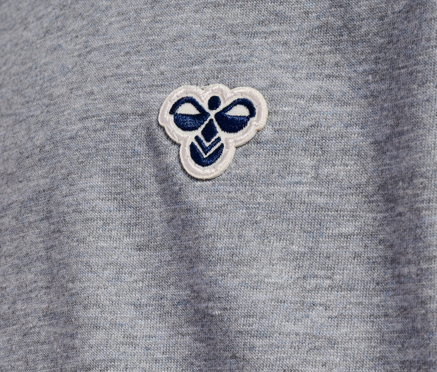 Gros plan sur un tissu gris clair avec un petit logo Hummel blanc et bleu cousu.