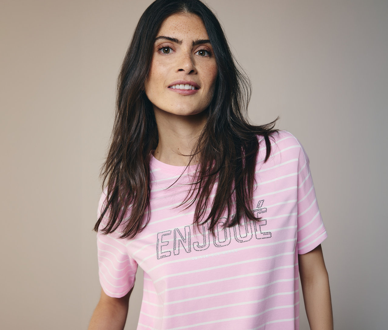 Une femme aux longs cheveux foncés porte un T-shirt CECIL à rayures roses et blanches avec le mot 'ENJOUE'.