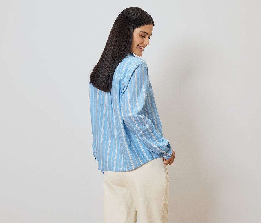 Une femme aux cheveux foncés, vue de dos, porte une chemise rayée bleu clair et un pantalon beige.