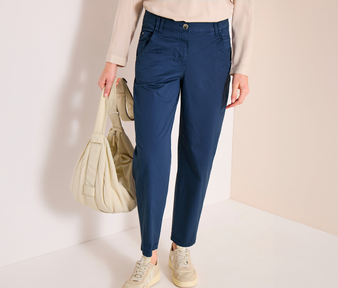 Une personne porte un pantalon barrel CECIL « Scarlett » bleu, un haut clair et des baskets claires, tenant un sac à bandoulière matelassé de couleur claire.