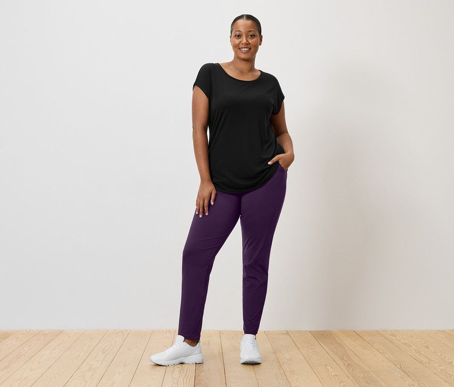 Femme portant un t-shirt noir, un pantalon violet et des baskets blanches debout devant un mur blanc.