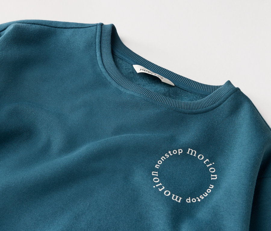 Gros plan d'un sweat-shirt bleu pétrole avec l'inscription "nonstop motion" en cercle.