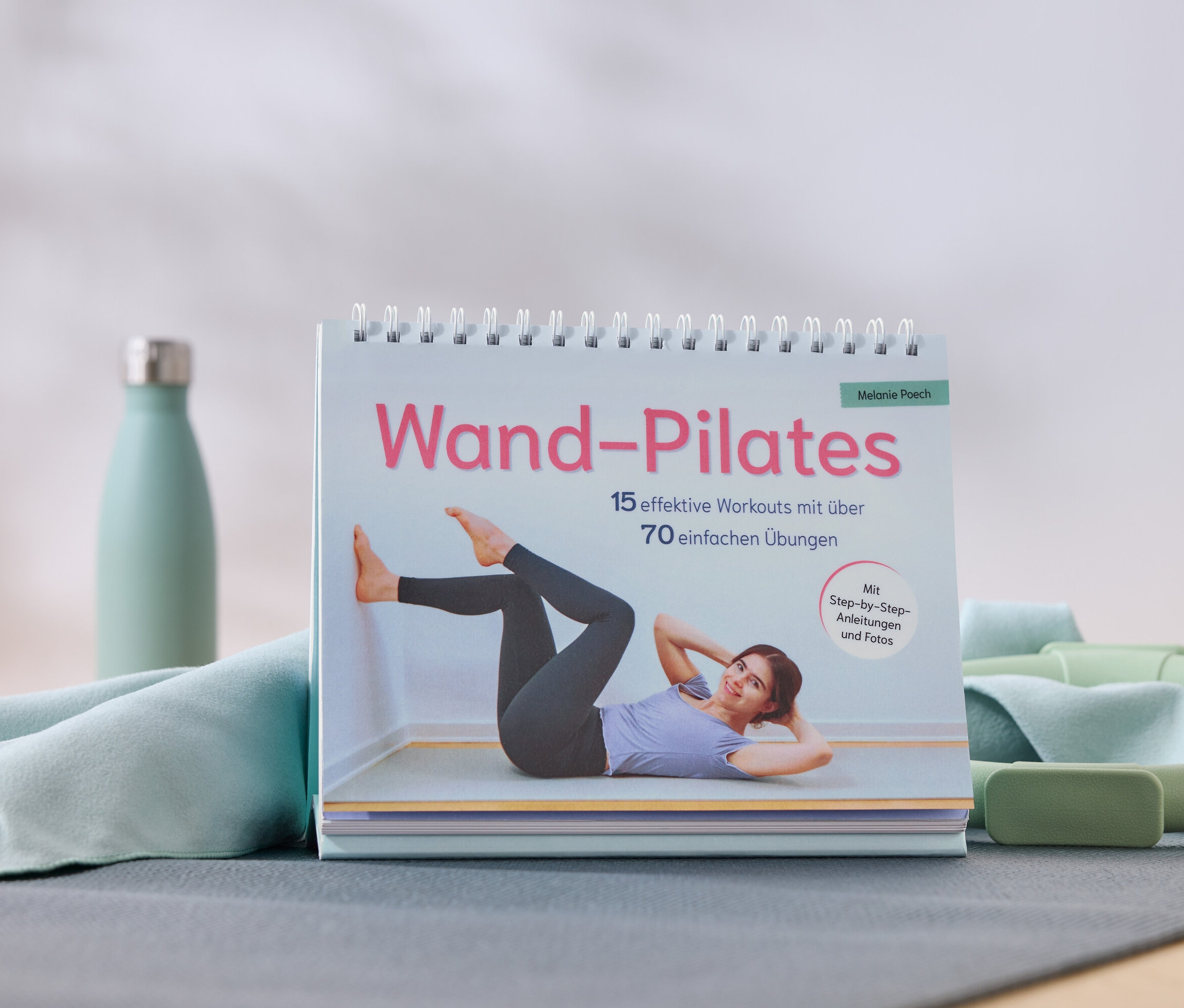 Un livre de Wall Pilates avec une femme faisant un exercice, à côté d'une bouteille d'eau, une serviette, un cercle de Pilates et un bloc.