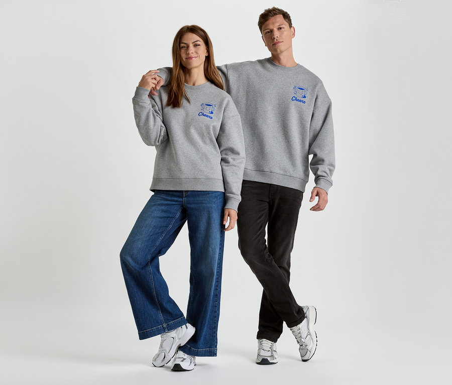 Une femme et un homme se tiennent côte à côte, portant des pulls gris avec des imprimés bleus "Cheers", combinés avec des jeans et des baskets.