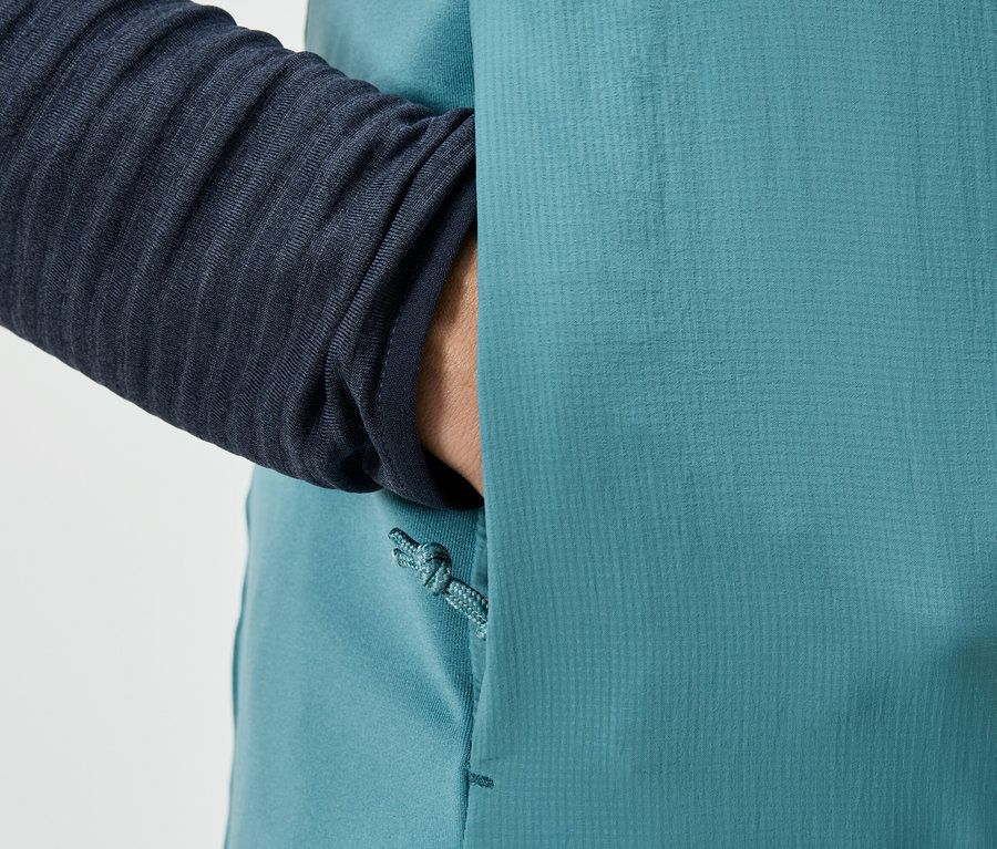Gros plan d'une personne qui met sa main dans la poche d'une veste turquoise, avec une manche bleu marine visible.