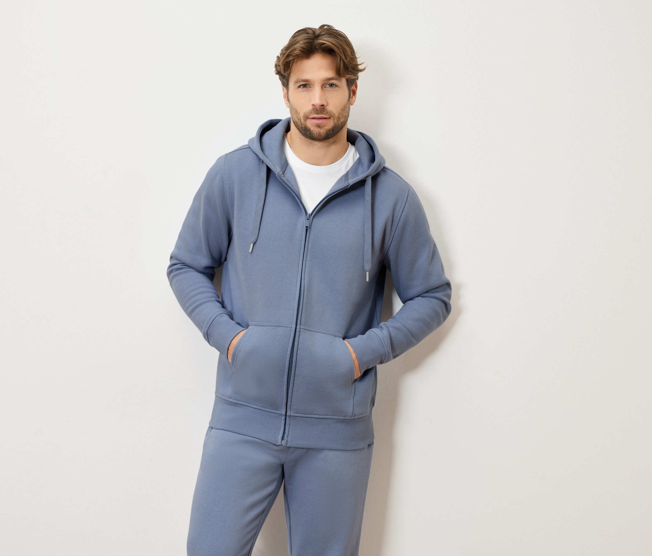 Un homme aux cheveux châtain clair portant un survêtement bleu à capuche et zippé et un t-shirt blanc, les deux mains dans les poches.