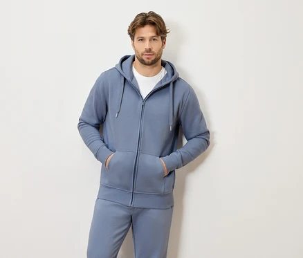 Un homme aux cheveux châtain clair portant un survêtement bleu à capuche et zippé et un t-shirt blanc, les deux mains dans les poches.