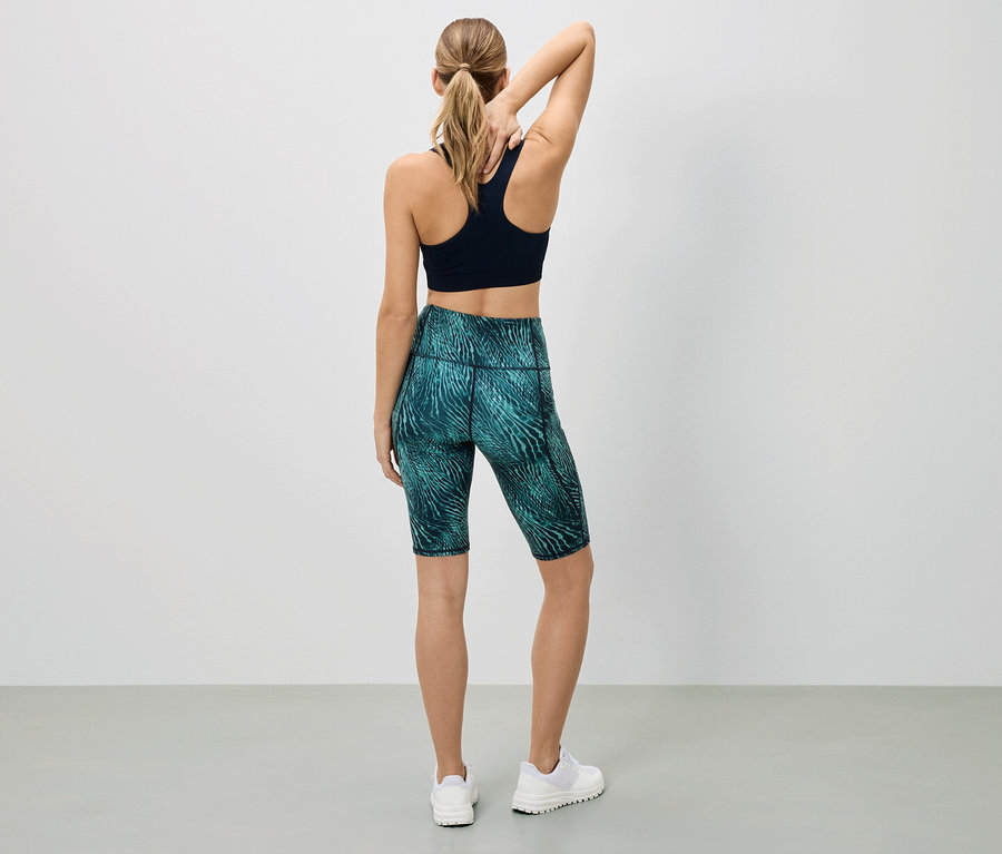 Une femme se tient dos à la caméra, portant un soutien-gorge de sport noir et un legging court à motifs.