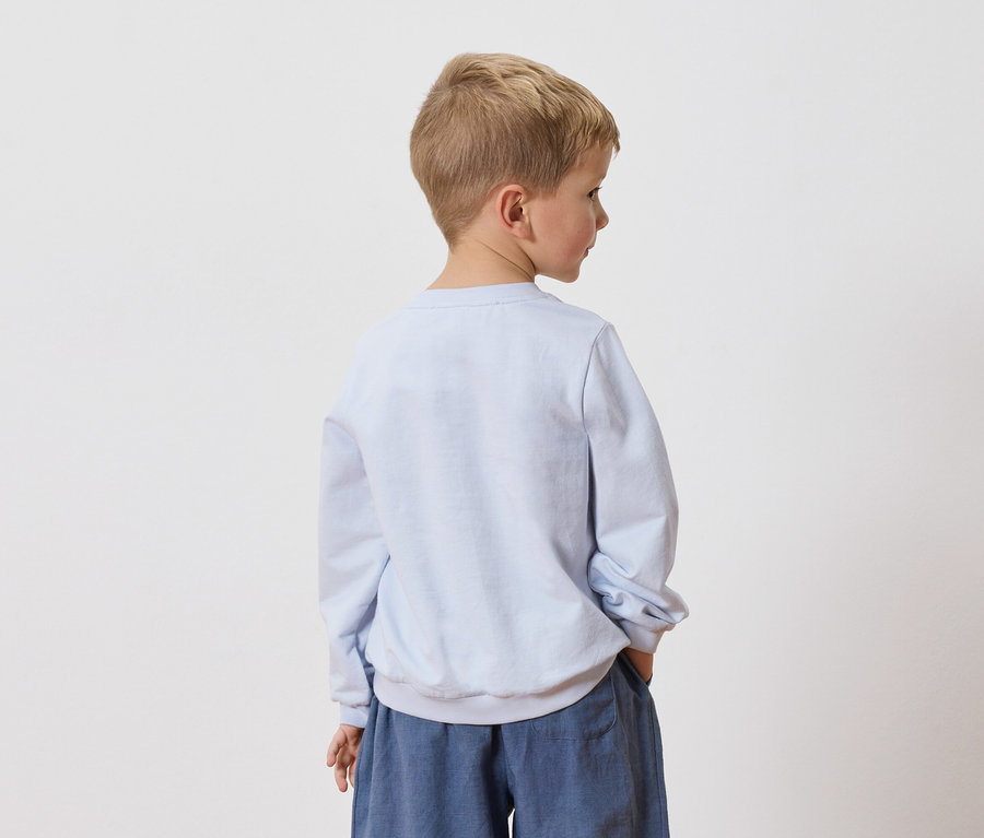 Un garçon blond se tient dos à la caméra, portant un sweat-shirt bleu clair et un pantalon bleu.