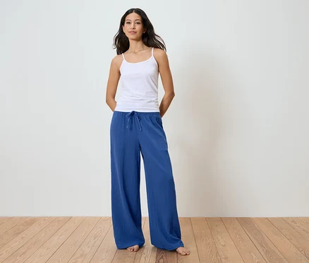 Femme debout pieds nus sur un plancher en bois, portant un débardeur blanc et un pantalon ample bleu avec cordon de serrage.