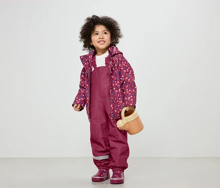 Une fillette aux cheveux bouclés porte une salopette bordeaux, une veste à fleurs et des bottes assorties, et tient un arrosoir.