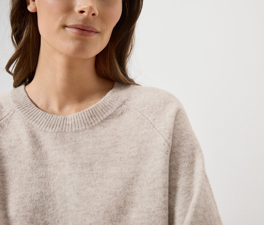 Une femme aux cheveux bruns porte un pull beige clair à col rond sur fond blanc.