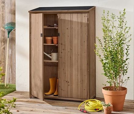 Une armoire en bois ouverte se trouve à l'extérieur avec des outils de jardinage, des pots, des bottes en caoutchouc et un arrosoir à l'intérieur.