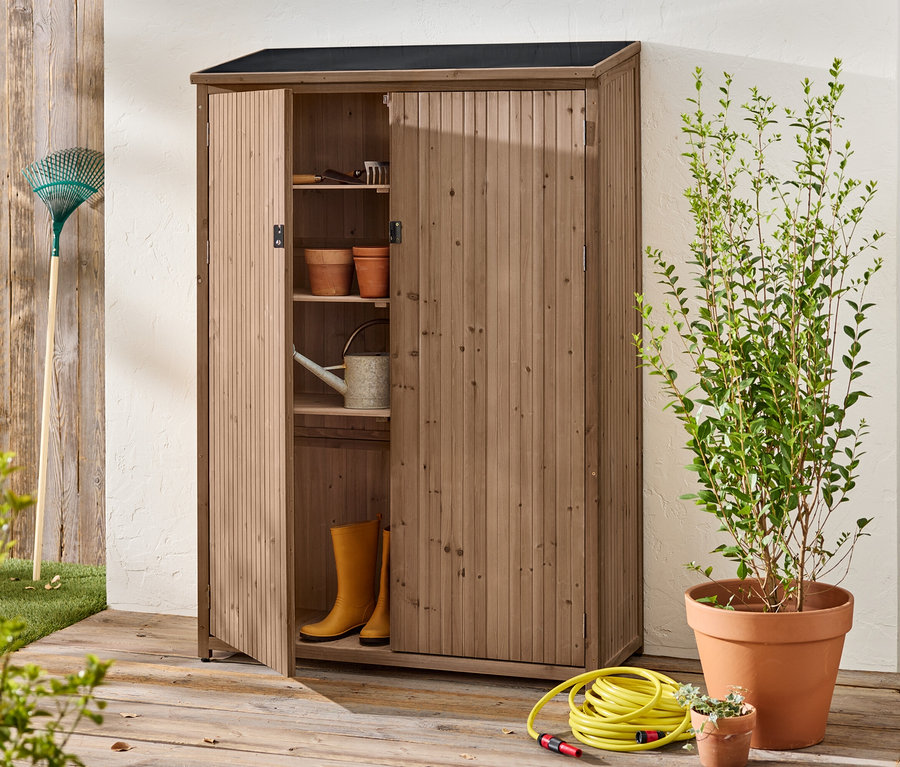 Une armoire en bois ouverte se trouve à l'extérieur avec des outils de jardinage, des pots, des bottes en caoutchouc et un arrosoir à l'intérieur.