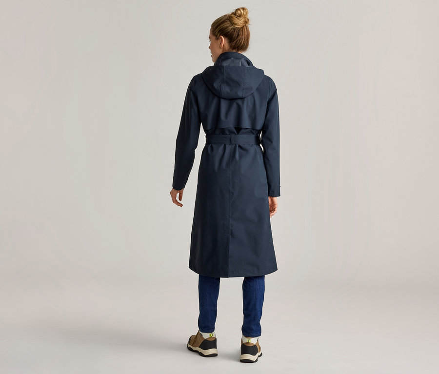 Vue arrière d'une femme portant un chignon, un long trench-coat bleu marine à capuche, un jean bleu et des chaussures marron.