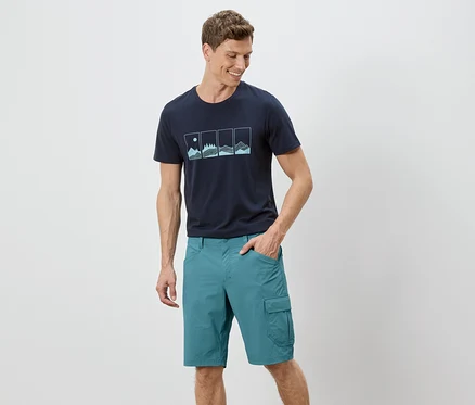 Un homme se tient debout avec une main dans sa poche devant un fond blanc. Il porte un t-shirt bleu marine avec un motif et un short vert.