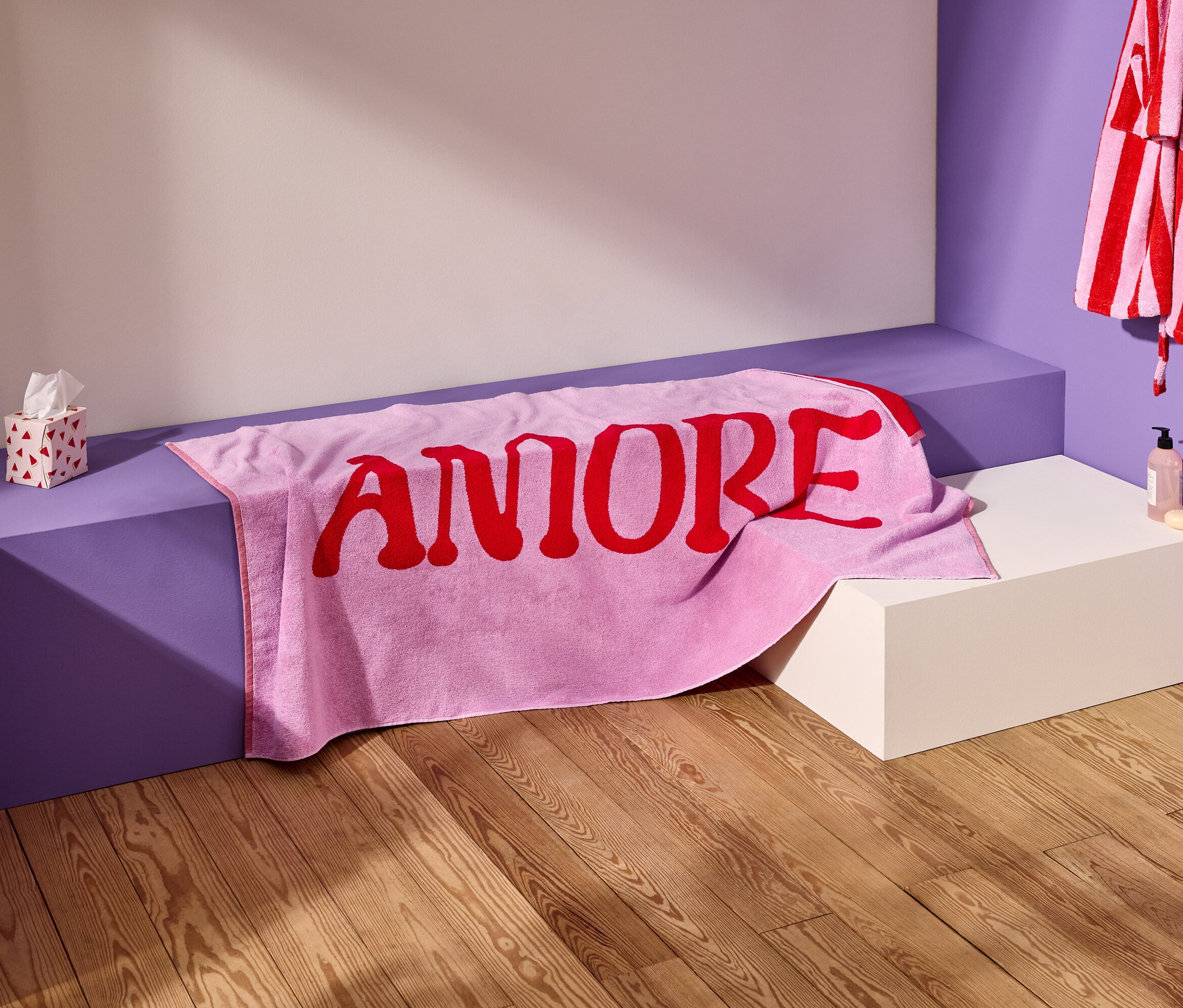 Une serviette rose avec le mot rouge "AMORE" est posée sur un banc violet. Un peignoir rayé est accroché au mur.