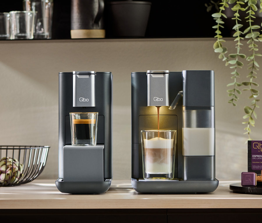 Deux machines à café Qbo MILK MASTER gris foncé sur un comptoir. L'une contient un espresso, l'autre prépare un latte macchiato. Des capsules de café sont à côté.