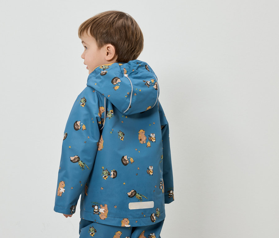 Petit garçon vu de dos portant un imperméable bleu à motifs d'ours et de hérissons sur fond blanc.