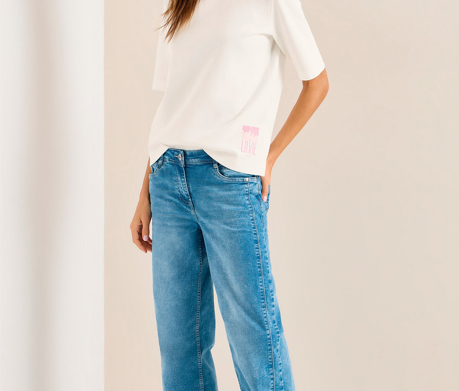 Femme souriante en sweatshirt CECIL blanc à motif rose, jean bleu, baskets blanches et casquette violet clair.