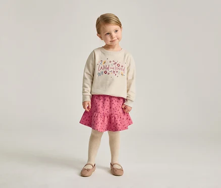 Une fillette blonde porte un sweat-shirt beige avec l'inscription "Wild and loved", une jupe rose et des ballerines.