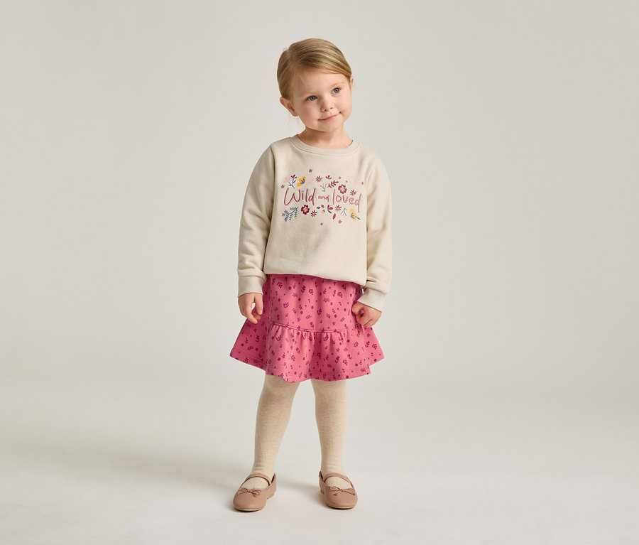 Une fillette blonde porte un sweat-shirt beige avec l'inscription "Wild and loved", une jupe rose et des ballerines.