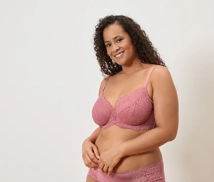 Une femme souriante aux cheveux bouclés porte un soutien-gorge en dentelle rose et une culotte assortie devant un fond uni.