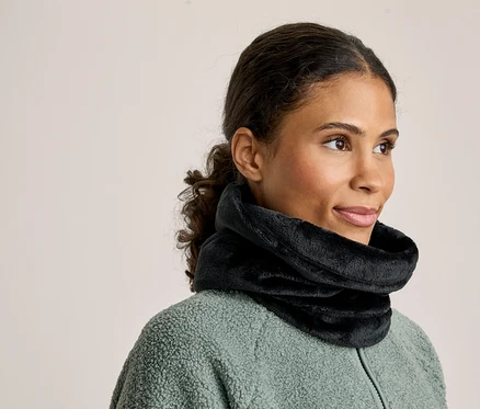 Une femme aux cheveux foncés attachés en arrière porte un cache-cou noir moelleux et une veste polaire vert clair, regardant vers sa droite.