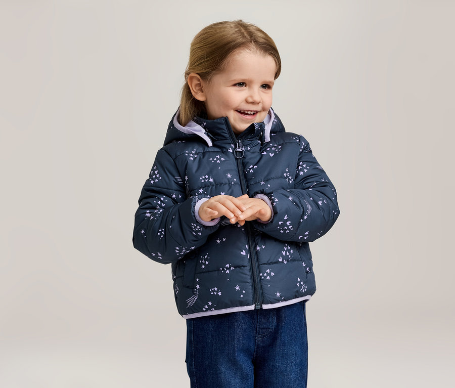 Une fillette souriante aux cheveux blonds porte une veste matelassée bleu marine à capuche avec un motif étoilé et un jean bleu.