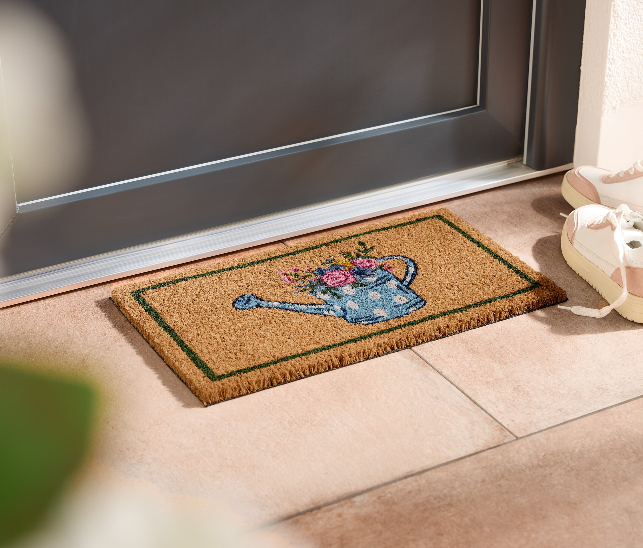 Un paillasson avec un motif d'arrosoir est placé devant une porte, à côté de chaussures de sport blanches.