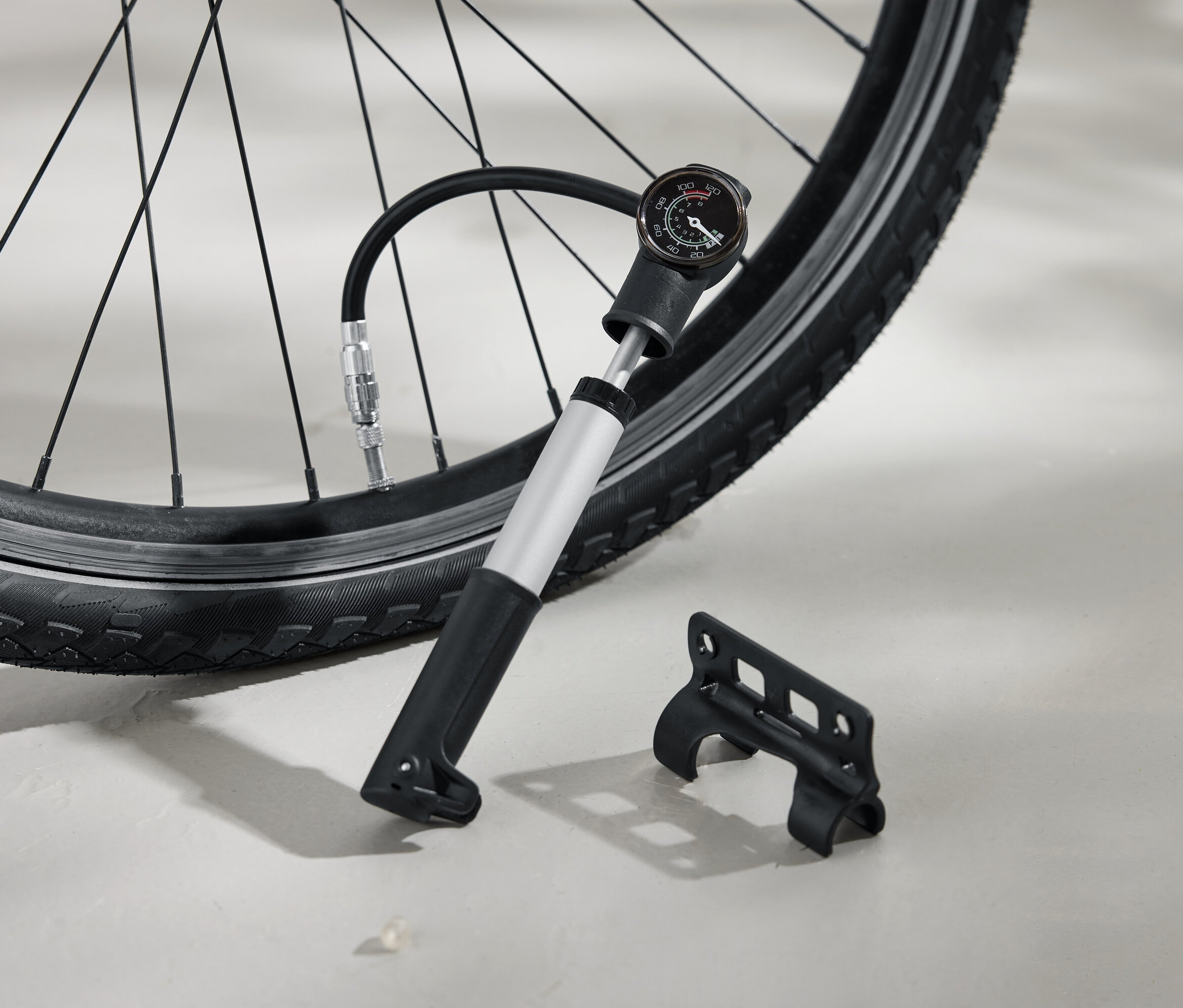Un pneu de vélo est gonflé avec une pompe à air argentée dotée d'une poignée noire et d'un manomètre analogique. Un support noir est posé sur le sol à côté.
