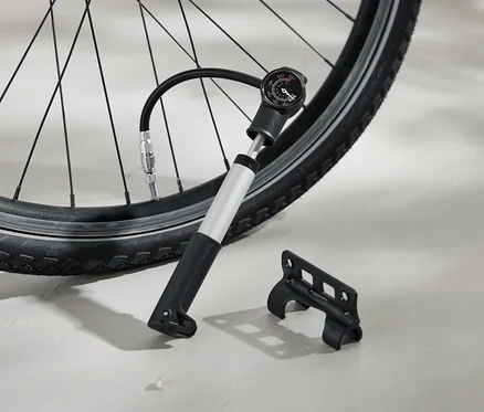 Un pneu de vélo est gonflé avec une pompe à air argentée dotée d'une poignée noire et d'un manomètre analogique. Un support noir est posé sur le sol à côté.