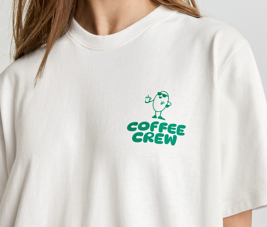 Gros plan d'une personne portant un t-shirt clair avec l'inscription verte "Coffee Crew" et un personnage de grain de café dessiné.