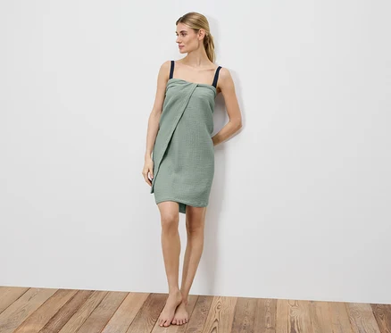 Une femme se tient pieds nus contre un mur blanc, enveloppée dans une robe en mousseline verte avec des bretelles foncées.