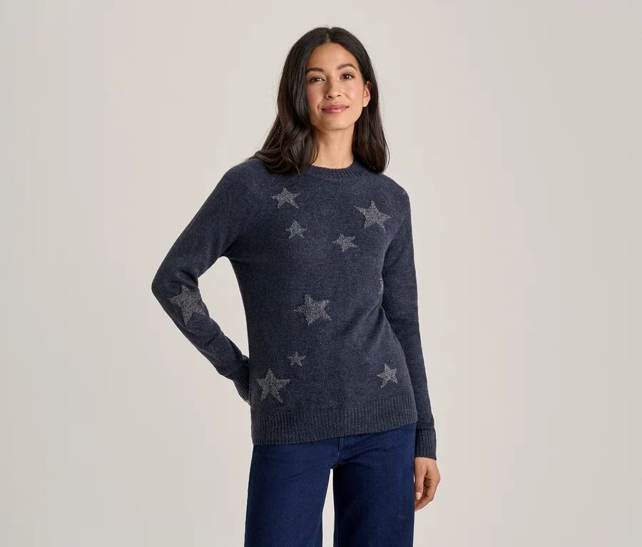 Une femme souriante se tient debout, les mains sur les hanches, vêtue d'un pull bleu foncé orné d'étoiles argentées et d'un jean bleu foncé.