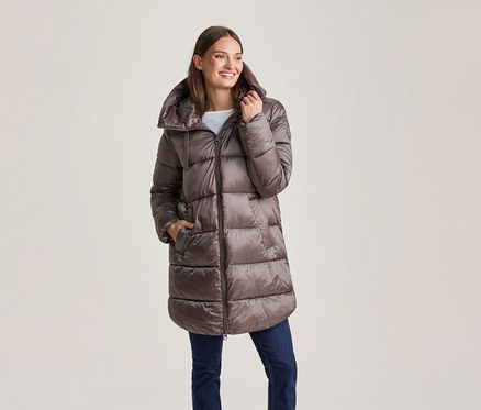 Femme souriante portant une longue doudoune marron brillante et un jean bleu, une main dans la poche.