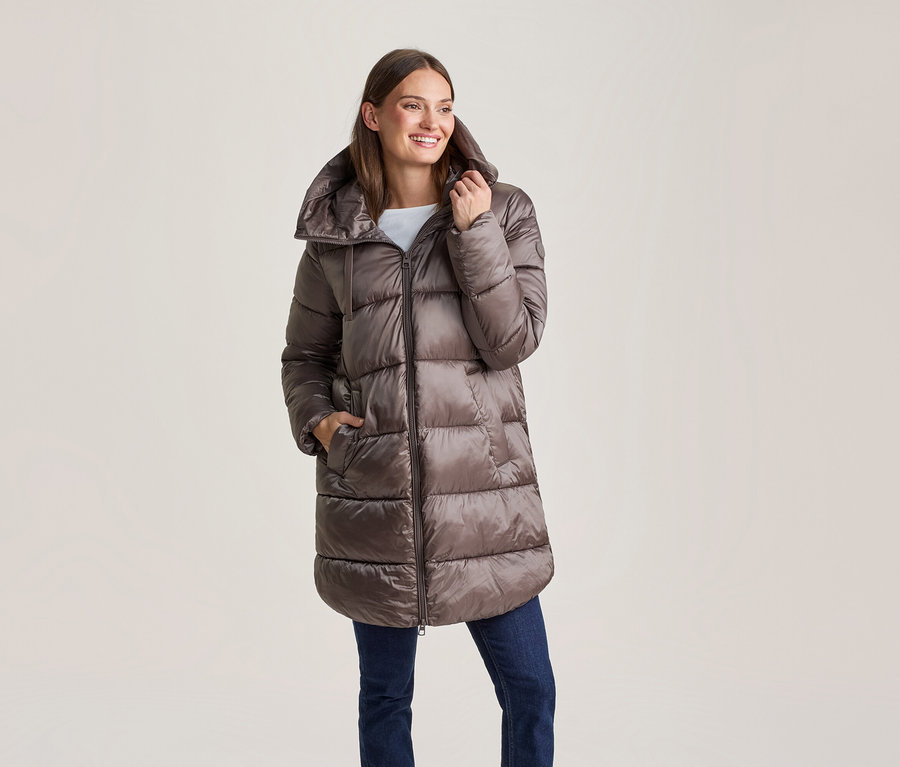 Femme souriante portant une longue doudoune marron brillante et un jean bleu, une main dans la poche.