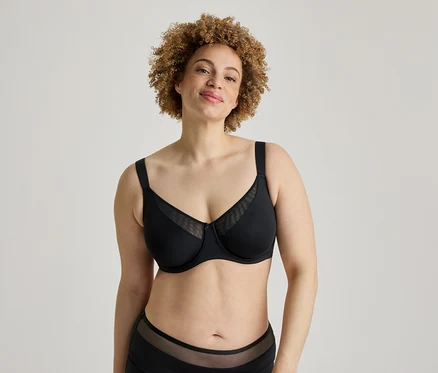 Une femme souriante aux cheveux bouclés se tient devant un fond blanc, vêtue d'un soutien-gorge noir avec des empiècements en maille et d'un slip noir assorti avec des détails en maille.