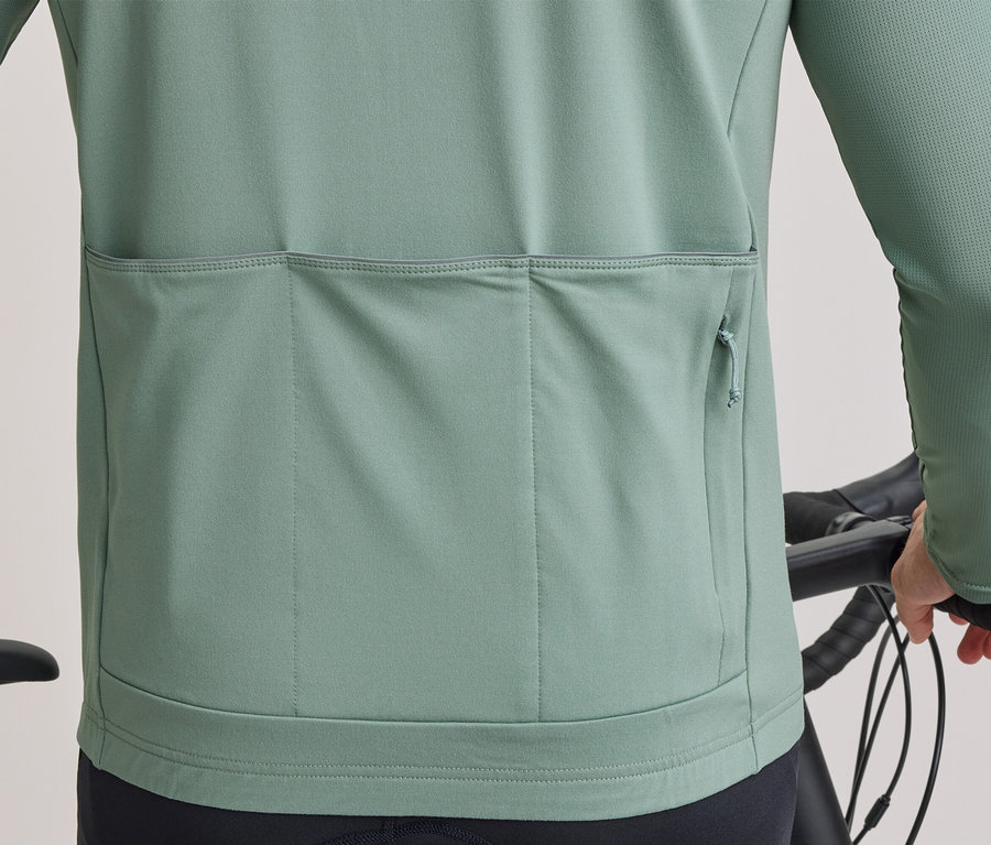 Dos d'une personne en maillot de cyclisme vert clair à manches longues, trois poches arrière dont une zippée, tenant un guidon de vélo.