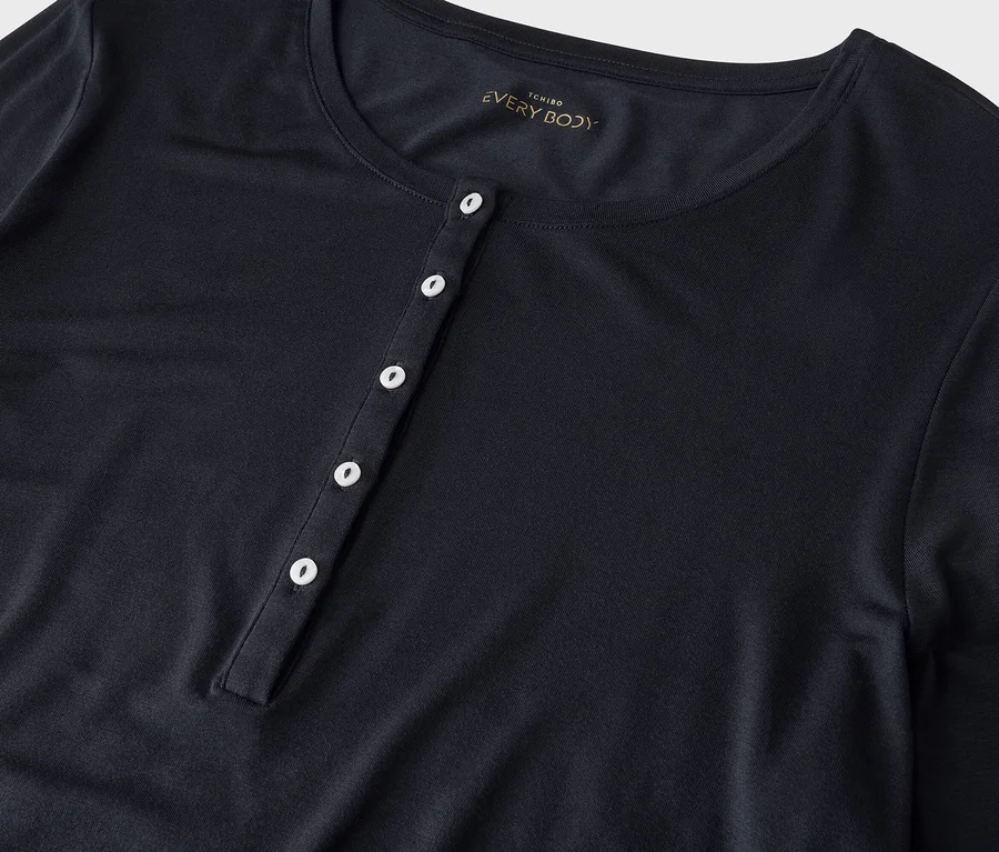 Un t-shirt à manches longues bleu foncé avec un col rond et une patte de boutonnage à cinq boutons blancs. L'étiquette "TCHIBO EVERY BODY" est visible.