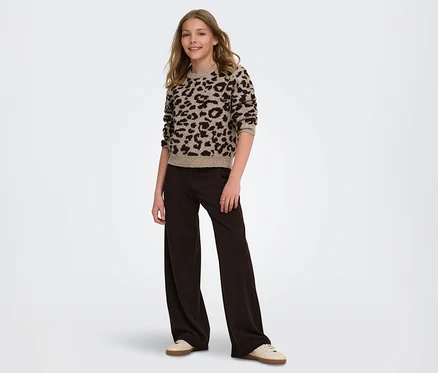 Une jeune fille souriante est debout, vêtue d'un pull à imprimé léopard beige et marron, d'un pantalon large marron foncé et de baskets claires.