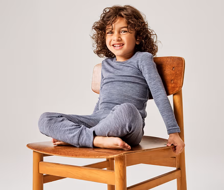 Un jeune enfant aux cheveux bouclés, vêtu d'un haut gris côtelé à manches longues et d'un pantalon assorti, est assis en tailleur sur une chaise en bois et sourit à l'appareil photo.