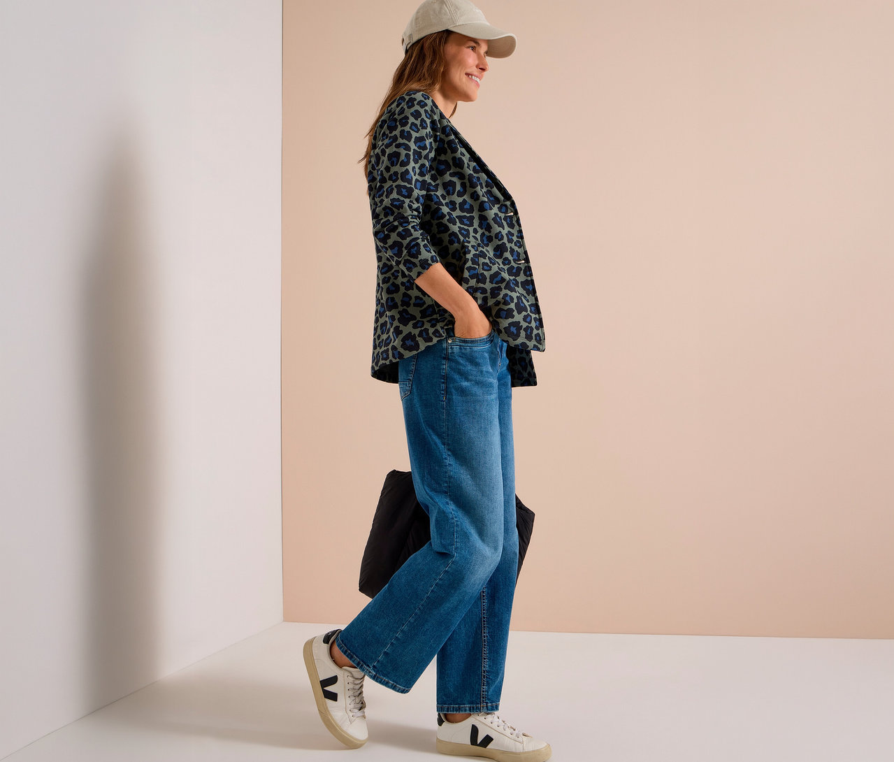Femme portant une casquette beige, un blazer en jersey à imprimé léopard vert et bleu, un jean large et des baskets blanches, debout, une main dans la poche et tenant un sac noir.