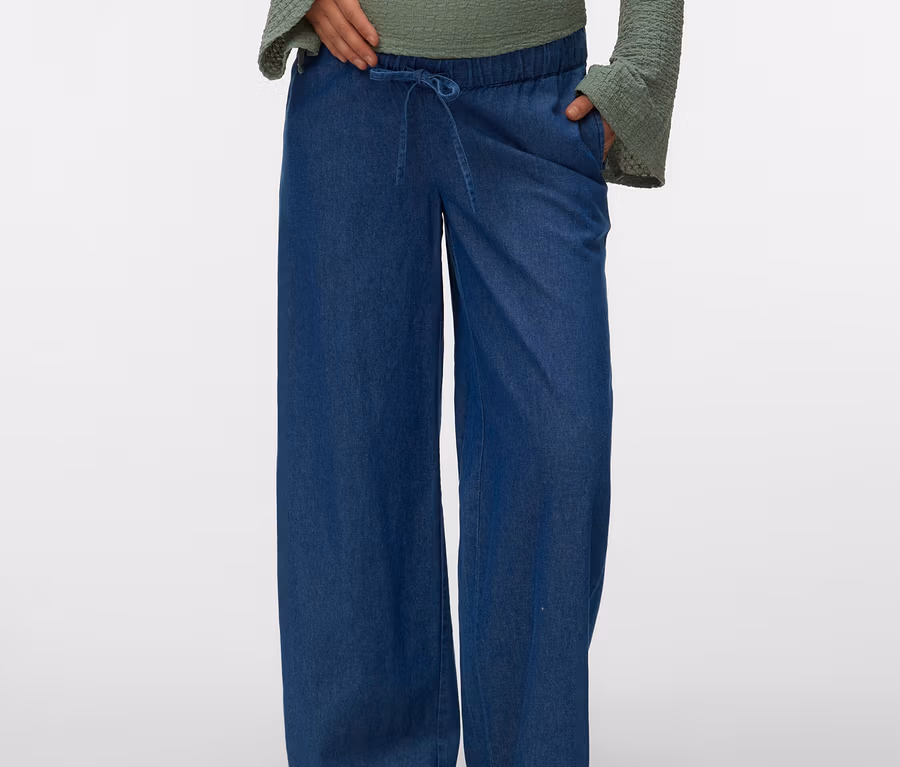 Une personne enceinte, la main sur le ventre, porte un chemisier vert à manches longues et un Jeans ample MAMALICIOUS « MLJAMAICA » bleu avec cordon de serrage, ainsi que des mules marron.