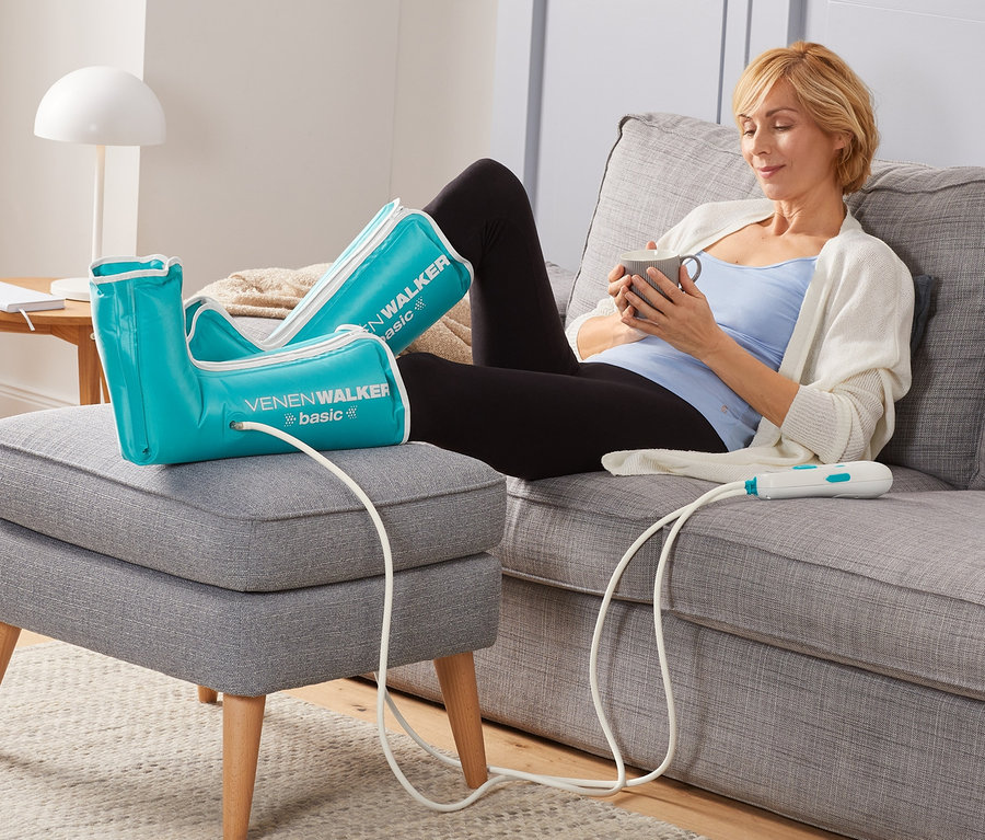 Une femme est assise sur un canapé et utilise l'appareil de thérapie « VenenWalker Basic » de VITALmaxx tout en buvant du café.