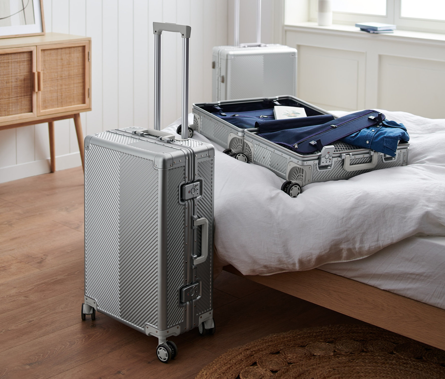 Sur un plancher en bois se trouve une petite valise en aluminium. Sur le lit se trouve une valise en aluminium moyenne ouverte et à l'arrière-plan, on peut voir une grande valise en aluminium.