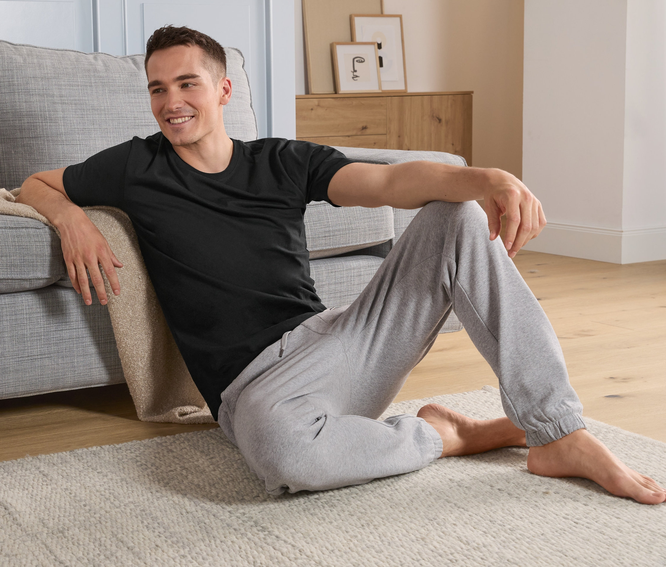 Un homme est assis sur le sol à côté d’un canapé et porte un t-shirt noir à encolure ronde.