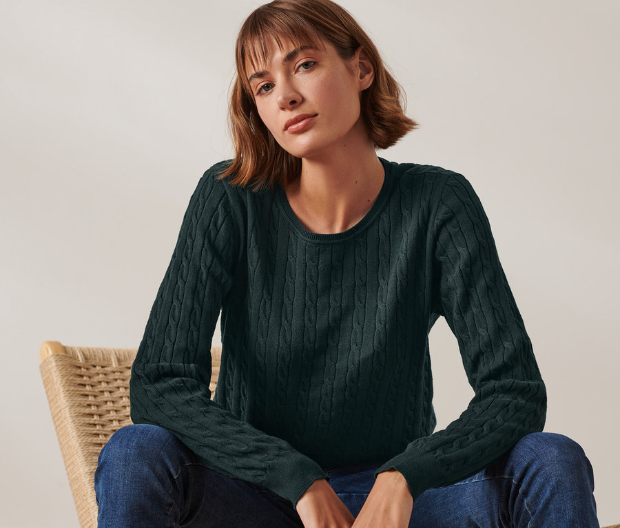 Une femme est assise sur une chaise en osier et porte un pull en maille torsadée vert foncé.